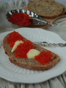 A slice of rustic bread topped with butter and red caviar on a white plate.